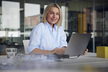 Lächelnde Geschäftsfrau mit Laptop sitzt am Schreibtisch im Büro - RBF08802