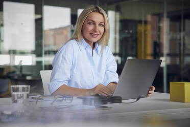 Smiling businesswoman with laptop sitting at desk in office - RBF08802