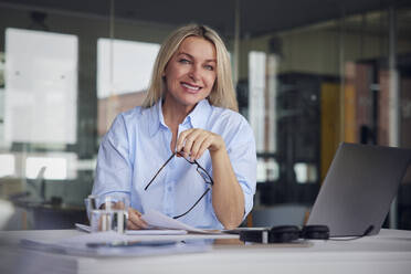 Smiling businesswoman holding paper and eyeglasses sitting by laptop at desk in office - RBF08803