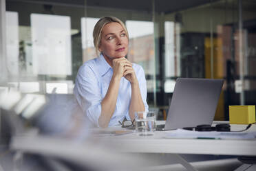 Thoughtful businesswoman with hand on chin sitting by laptop at desk in office - RBF08805