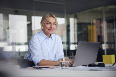Smiling businesswoman with laptop at desk in office - RBF08807