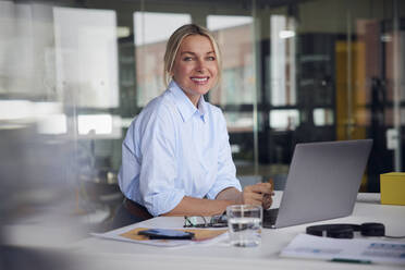 Happy businesswoman with laptop sitting at desk in office - RBF08808