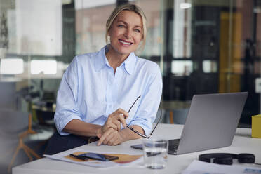 Smiling businesswoman holding eyeglasses and laptop at desk in office - RBF08809