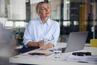 Businesswoman holding eyeglasses and laptop at desk in office - RBF08811