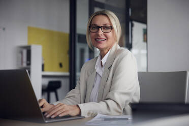 Lächelnde Geschäftsfrau mit Brille und Laptop am Schreibtisch im Büro - RBF08827