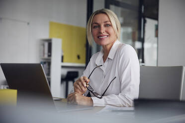 Glückliche Geschäftsfrau mit Brille und Laptop am Schreibtisch im Büro - RBF08828