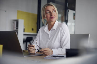 Businesswoman holding eyeglasses with laptop at desk in office - RBF08829