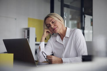 Geschäftsfrau mit geschlossenen Augen, die eine Brille mit Laptop am Schreibtisch im Büro hält - RBF08830