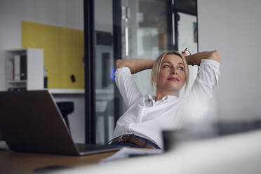 Businesswoman sitting with hands behind head with laptop at desk - RBF08831