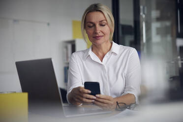 Smiling businesswoman using smart phone and laptop at desk in office - RBF08836