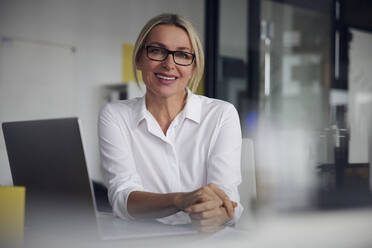 Lächelnde Geschäftsfrau mit Brille sitzt mit Laptop am Schreibtisch im Büro - RBF08838