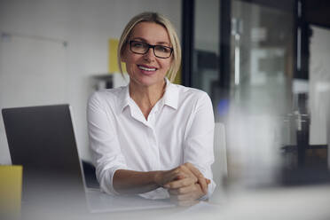 Smiling businesswoman wearing eyeglasses sitting with laptop at desk in office - RBF08838