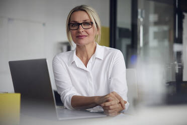 Geschäftsfrau mit Brille sitzt mit Laptop am Schreibtisch im Büro - RBF08839