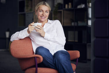 Lächelnde Geschäftsfrau mit Kaffeetasse auf einem Stuhl im Büro sitzend - RBF08841
