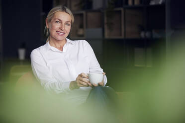 Glückliche Geschäftsfrau mit Kaffeetasse auf einem Stuhl im Büro sitzend - RBF08851