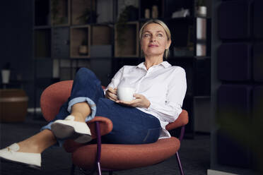 Geschäftsfrau mit Kaffeetasse, die mit übereinander geschlagenen Beinen auf einem Stuhl sitzt - RBF08858