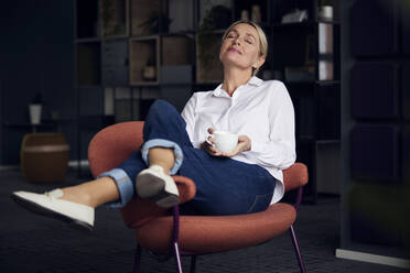 Geschäftsfrau mit Kaffeetasse sitzt mit geschlossenen Augen auf einem Stuhl im Büro - RBF08859