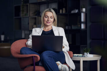 Businesswoman using laptop sitting on chair in office - RBF08867
