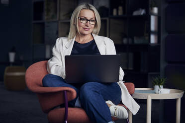 Lächelnde Geschäftsfrau mit Laptop auf einem Stuhl im Büro sitzend - RBF08869