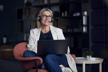 Smiling businesswoman wearing headset working on laptop at office - RBF08870