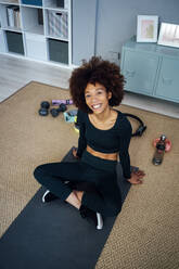 Glückliche junge Frau mit Afrofrisur sitzt auf Übungsmatte zu Hause - GIOF15047