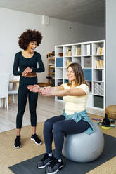 Young woman teaching friend exercising sitting on fitness ball in living room - GIOF15061