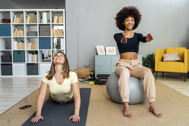 Happy young woman sitting on fitness ball by woman doing exercise in living room at home - GIOF15091