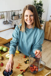 Happy blond woman with fresh fruits in blender standing at table in kitchen - GIOF15108