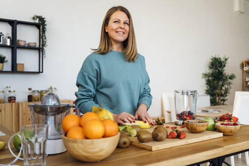 Smiling blond woman with fresh fruits at table standing in kitchen - GIOF15109