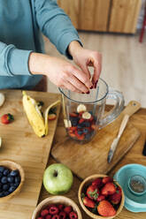 Woman putting fresh chopped fruits in blender at table in kitchen - GIOF15110