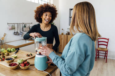 Happy young woman blending smoothie talking with friend in kitchen - GIOF15113