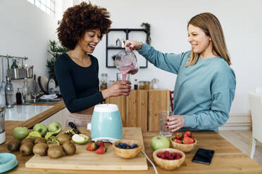 Lächelnde blonde Frau schenkt einer Freundin in der Küche zu Hause einen Smoothie ein - GIOF15116