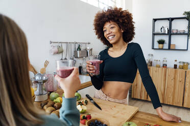 Glückliche junge Frau hält ein Glas Smoothie und unterhält sich mit einem Freund in der Küche - GIOF15120