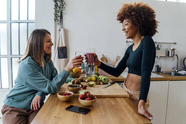 Happy young woman toasting glass of smoothie with friend sitting at table in kitchen - GIOF15121