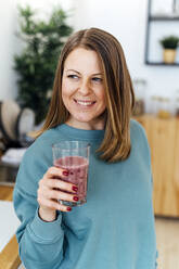Happy woman holding glass of smoothie in kitchen at home - GIOF15126