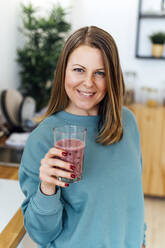 Smiling woman with glass of smoothie in kitchen at home - GIOF15127