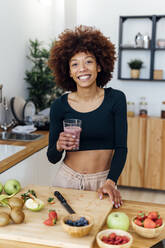 Happy young woman with glass of smoothie standing in kitchen at home - GIOF15128