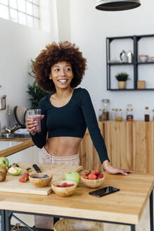 Glückliche junge Frau mit einem Glas Smoothie am Tisch in der Küche stehend - GIOF15129