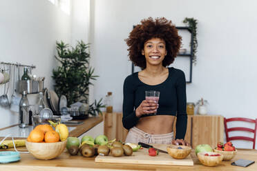 Happy young woman with Afro hairstyle holding glass of smoothie standing at table in kitchen - GIOF15130