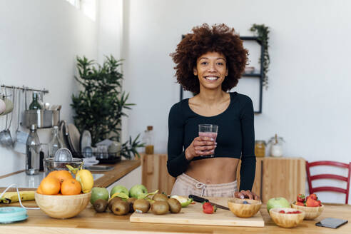 Glückliche junge Frau mit Afrofrisur hält ein Glas Smoothie und steht am Tisch in der Küche - GIOF15130