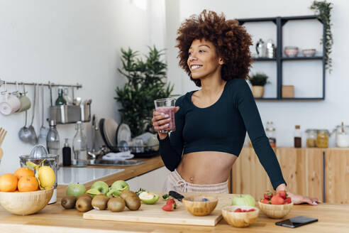Smiling woman with Afro hairstyle holding glass of smoothie standing in kitchen - GIOF15131
