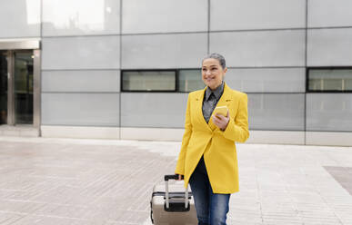 Smiling businesswoman holding smart phone walking with wheeled luggage on footpath - JCCMF05883