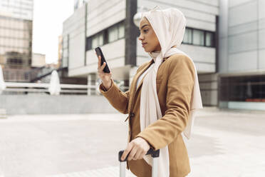 Businesswoman wearing hijab surfing net through smart phone standing outside modern building - JCCMF05888