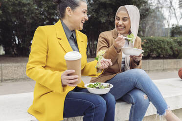 Cheerful business colleagues with salad bowls and disposable coffee cup sitting on steps - JCCMF05899