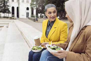 Businesswoman holding salad bowl and disposable coffee cup talking with colleague sitting on steps - JCCMF05900