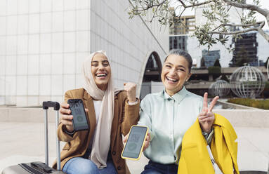 Happy businesswomen gesturing showing vaccination QR certificate on smart phones - JCCMF05906