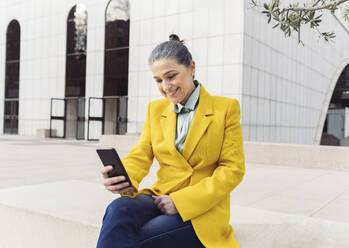 Glückliche Geschäftsfrau, die auf einer Bank sitzend mit ihrem Smartphone im Internet surft - JCCMF05908