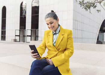 Happy businesswoman surfing net through smart phone sitting on bench - JCCMF05908