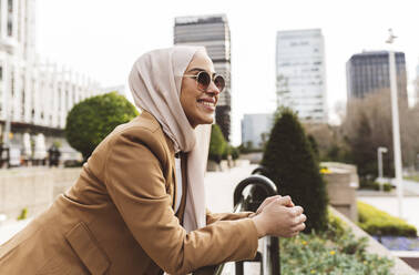 Glückliche Frau mit Sonnenbrille, die sich am Geländer eines Büroparks anlehnt - JCCMF05940