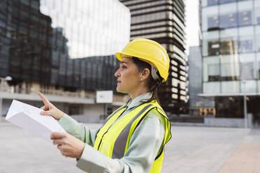 Ingenieur mit Schutzhelm und Blaupause bei der Arbeit außerhalb eines modernen Gebäudes - JCCMF05964